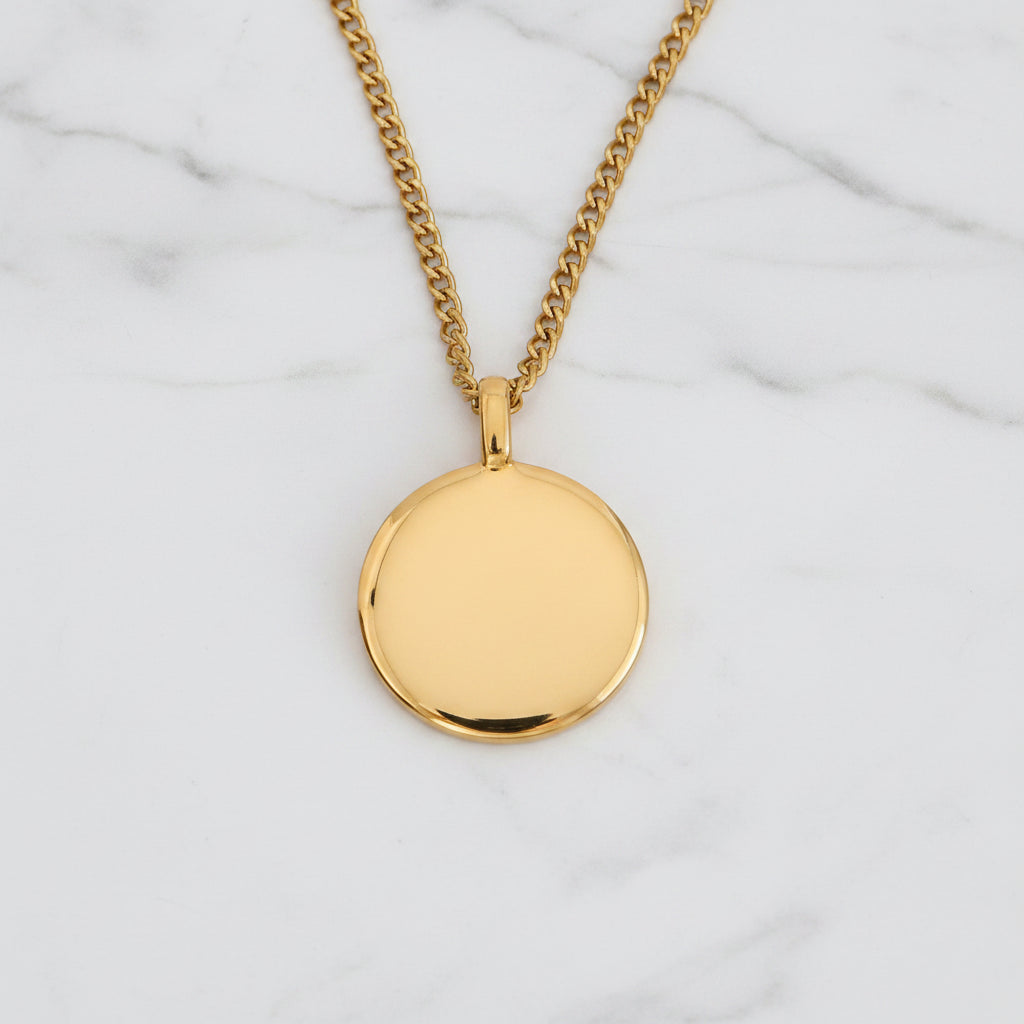 A gold pendant necklace with a flat, circular disc charm and a matching curb chain, displayed on a textured beige surface.