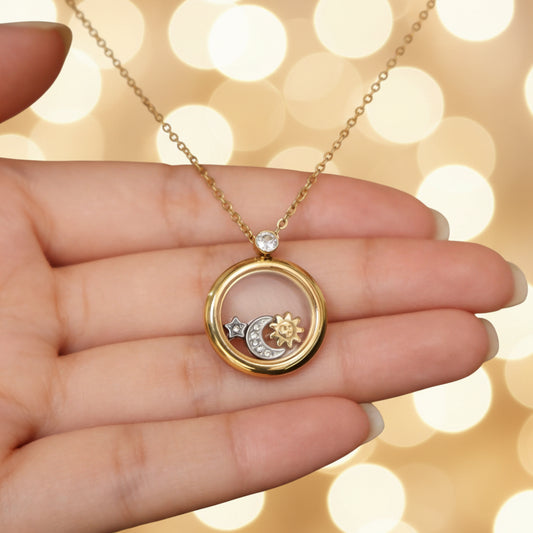 A gold-toned lugu pendant necklace with a crescent moon, sun, and small star design, held in a person's hand. The necklace features a gold chain and a circular pendant with a gold-toned ring, a small clear gem, and celestial accents.
