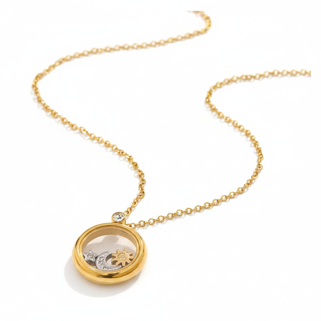 A gold chain necklace with a round locket pendant resting on a textured beige surface. The pendant features a gold outer frame and a clear crystal window showing small gold and silver mechanical gears inside.