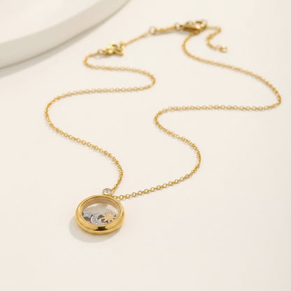 A gold chain necklace with a round locket pendant, displayed on a textured cream surface with a small stone prop in the corner.